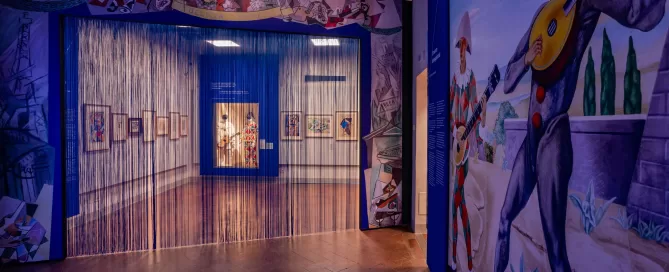 Veduta di una sala della collezione permanente dedicata a Gino Severini al MAEC. L'ingresso è incorniciato da decorazioni ispirate alla Commedia dell'arte e da un sipario di fili trasparenti. Sulla parete di destra è visibile una grande immagine con figure di Arlecchini musicisti, mentre sullo sfondo si intravedono opere esposte nelle sale. L'allestimento richiama l'universo teatrale e decorativo che caratterizza una parte importante della produzione di Severini.