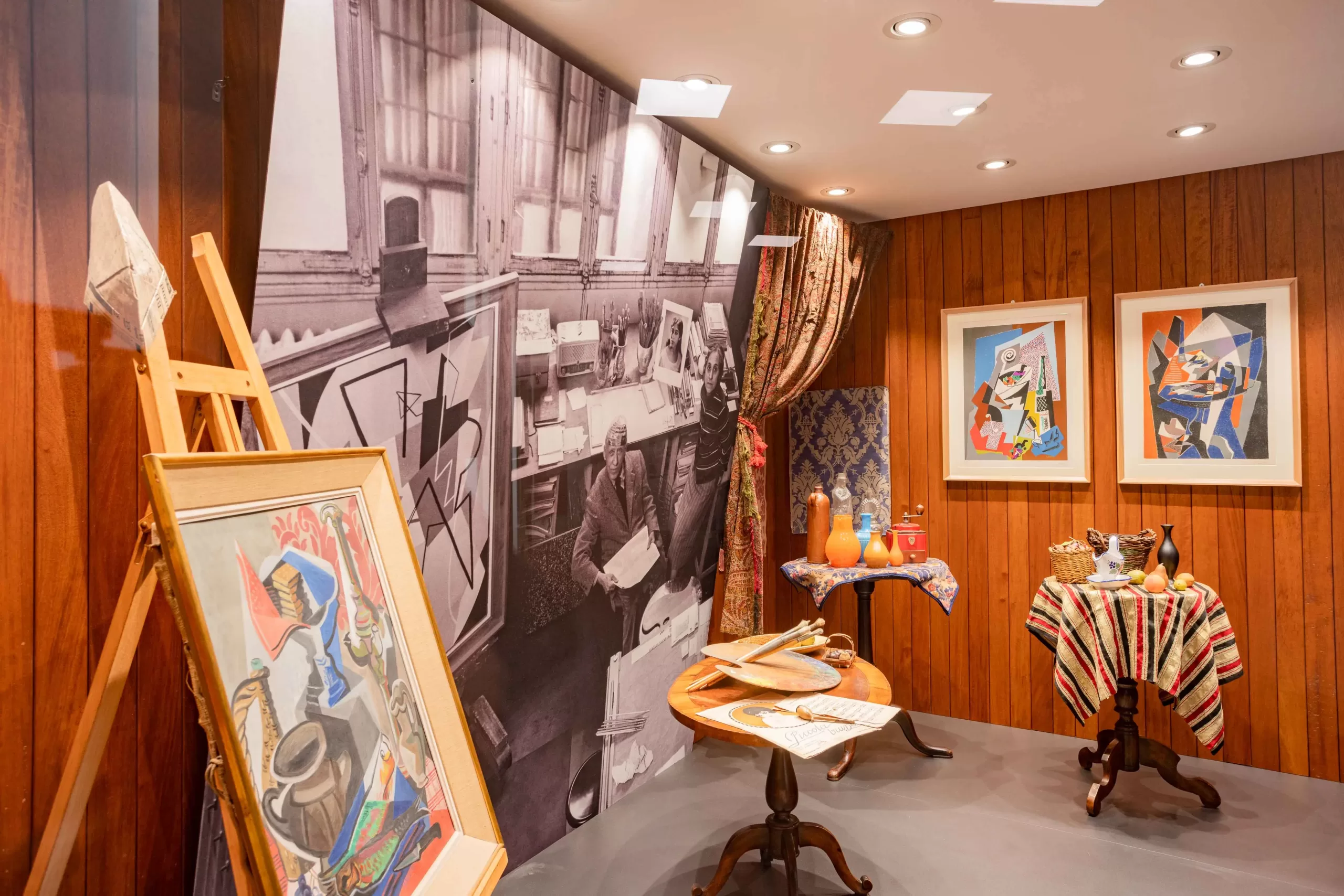 Ricostruzione dell'atelier parigino di Gino Severini al MAEC. La stanza presenta pareti rivestite in legno e una grande fotografia in bianco e nero dello studio dell'artista. Sono esposti dipinti, tavolini con oggetti decorativi, strumenti da lavoro e tessuti colorati che evocano l'ambiente creativo in cui Severini visse e lavorò a Parigi.