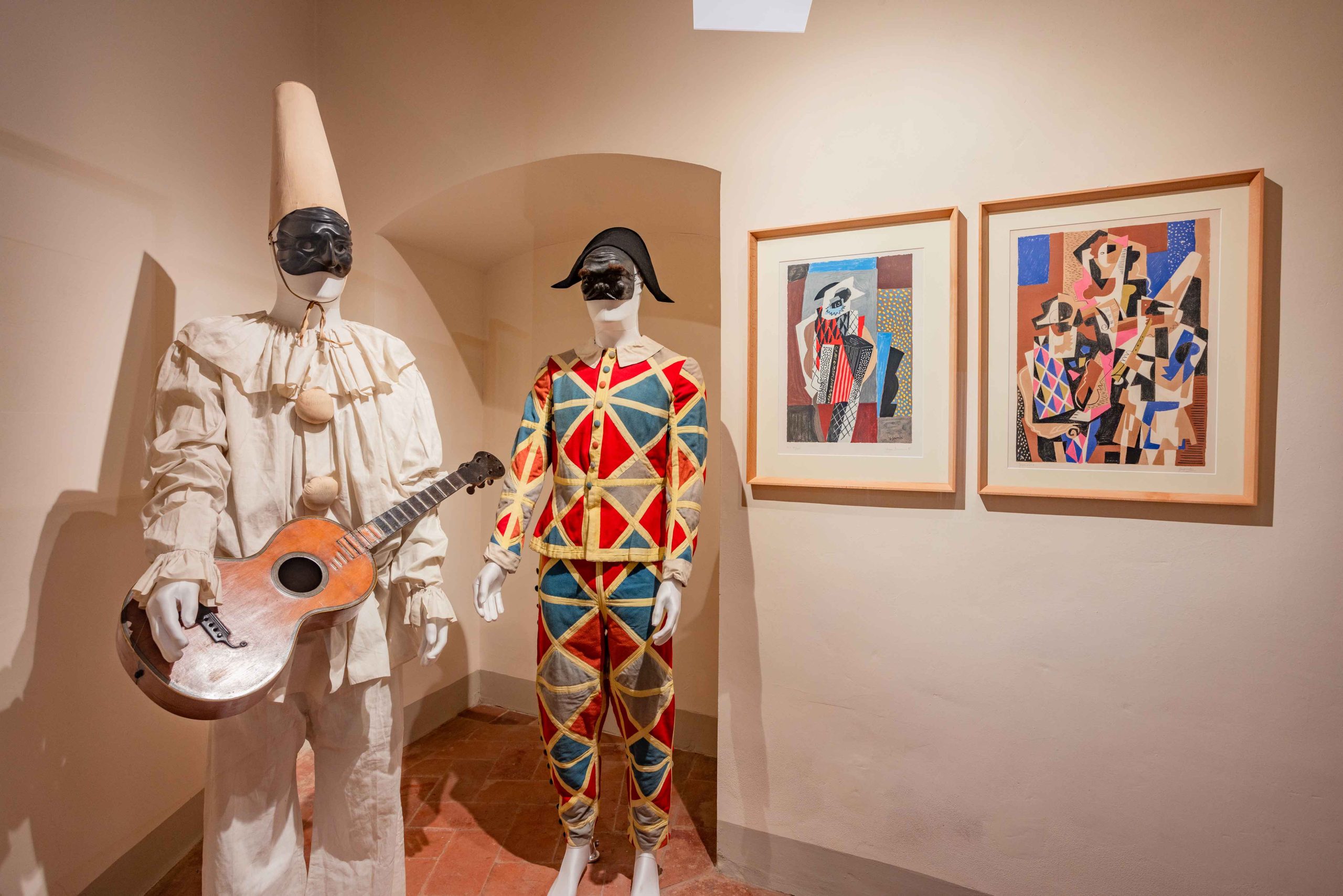Vetrina della collezione permanente dedicata a Gino Severini al MAEC. Due manichini indossano costumi ispirati ai personaggi della Commedia dell'arte: a sinistra un costume bianco con cappello a cono e una chitarra tra le mani, a destra un costume di Arlecchino con il tradizionale motivo a losanghe rosse, blu e gialle. Sulla parete accanto sono esposte due opere di Severini dedicate al tema delle maschere e del teatro.
