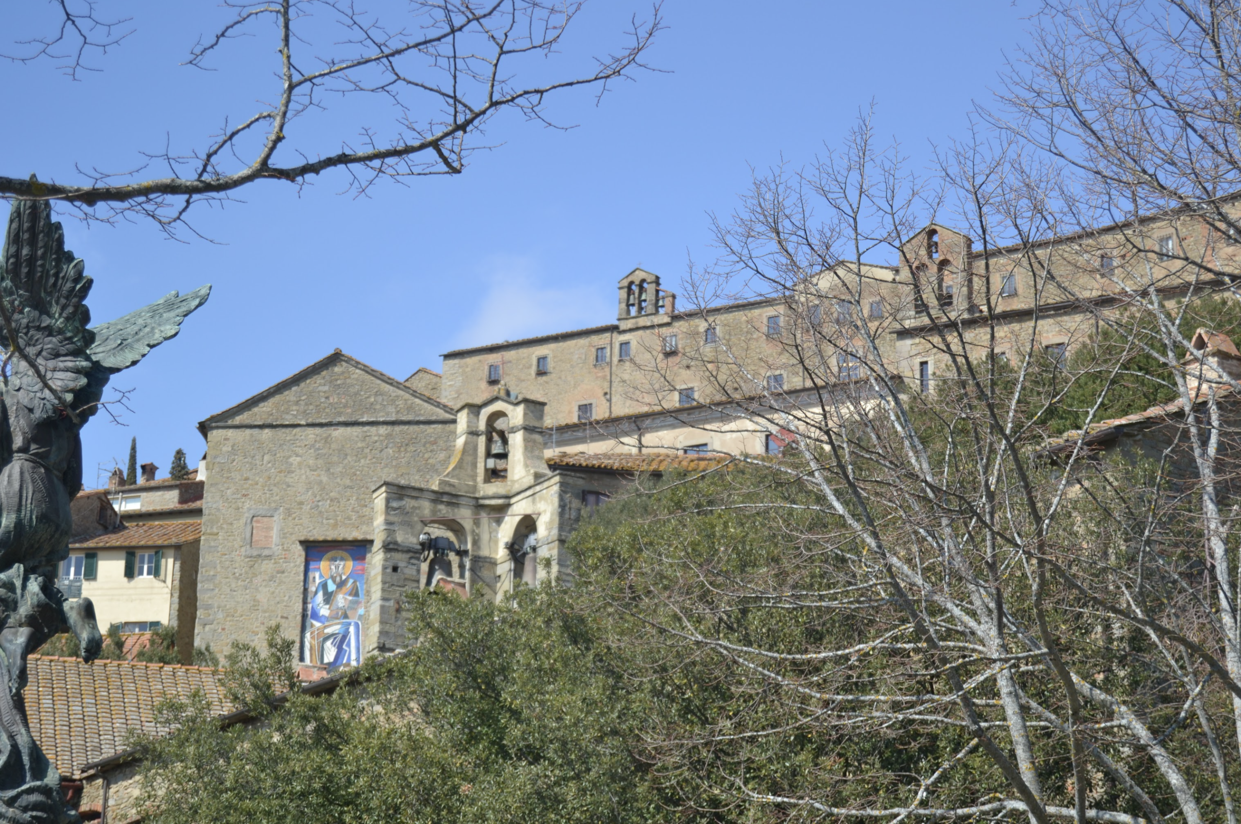 Vista panoramica del centro storico di Cortona. In primo piano si intravedono alberi e, sul lato sinistro, parte della statua alata dedicata all'eroe etrusco Dardano. Al centro si trova la chiesa di San Francesco, riconoscibile dalla facciata in pietra e dal piccolo campanile a vela. Sullo sfondo si sviluppa il complesso monumentale di Sant'Agostino, che domina il pendio con i suoi edifici in pietra. Il cielo è limpido e azzurro.